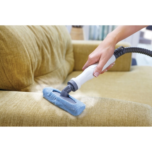 A hand using a BLACK+DECKER steam cleaning device with a blue microfiber attachment to clean a yellow upholstered sofa.