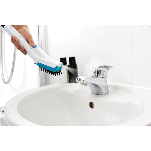 A person using a BLACK+DECKER steam cleaning brush to clean a bathroom sink faucet. The brush emits steam while cleaning the shiny chrome faucet.