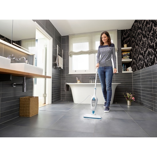 BLACKDECKER FSMH13E5 steam mop cleaning gray tile bathroom floor, held by person in striped shirt.