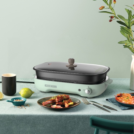BLACK+DECKER GMF1400 electric multi-cooker with glass lid on table, surrounded by dishes and a cup.