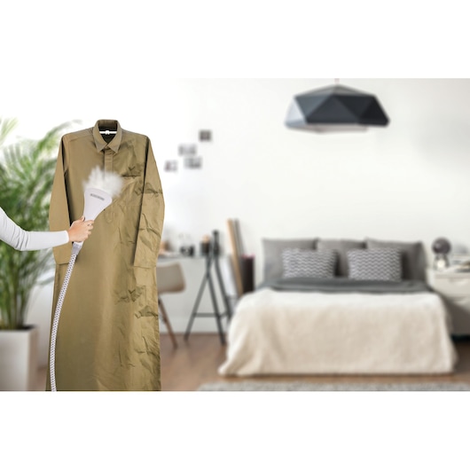 BLACKDECKER GSTM2050 garment steamer in use on a wrinkled shirt, bedroom background.