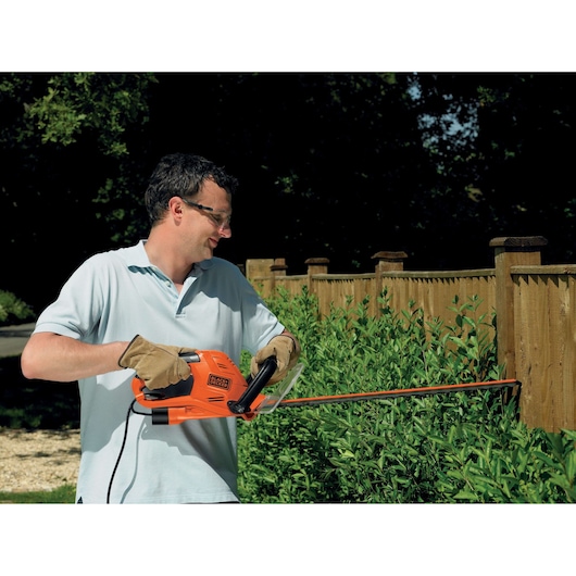 Photo of person using BLACK+DECKER GT5560 orange electric hedge trimmer to trim green bushes near wooden fence.