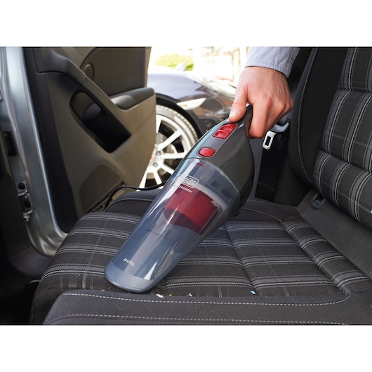 A handheld BLACK+DECKER vacuum cleaner is being used to clean the fabric seat of a car, operated by a person’s hand. The car door is open and another vehicle is visible outside.
