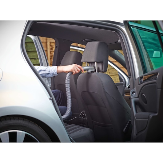 A person is using a handheld vacuum cleaner with a hose attachment to clean the headrest area of a car seat inside a vehicle.