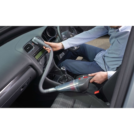 Person using a BLACK+DECKER handheld vacuum cleaner inside a car, cleaning the dashboard area with a flexible hose attachment.