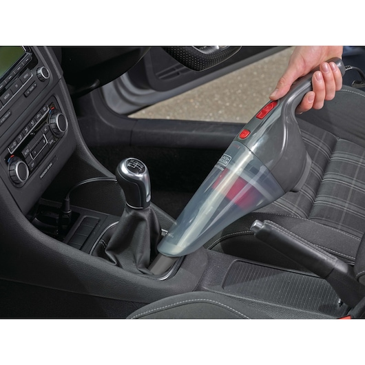 A hand holding a BLACK+DECKER handheld vacuum cleaner, cleaning the interior of a car near the gear shift and seat.