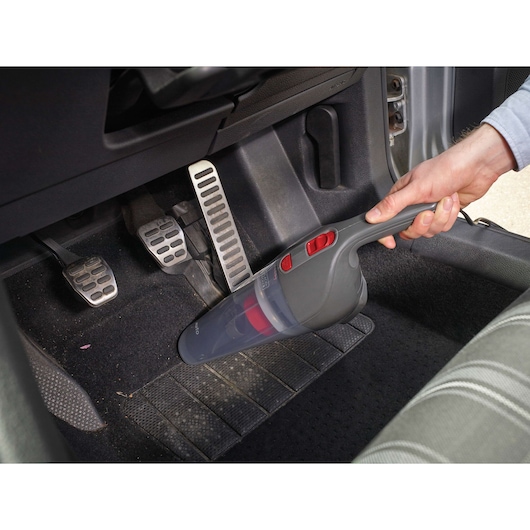 A person uses a BLACK+DECKER handheld vacuum cleaner to clean the floor area near the pedals inside a car.