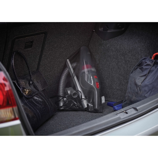 A BLACK+DECKER portable vacuum cleaner stored in the trunk of a car, placed in a mesh bag alongside a handbag and another bag, highlighting its compact storage for automotive use.