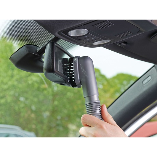 A hand uses a vacuum cleaner with a brush attachment to clean the area around a car's rear-view mirror and windshield.