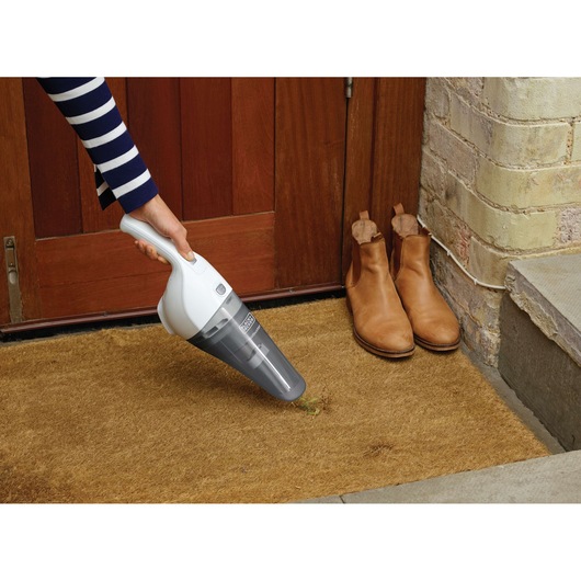 Photo of BLACKDECKER NVB215WN handheld vacuum cleaning a doormat near brown boots and a wooden door.