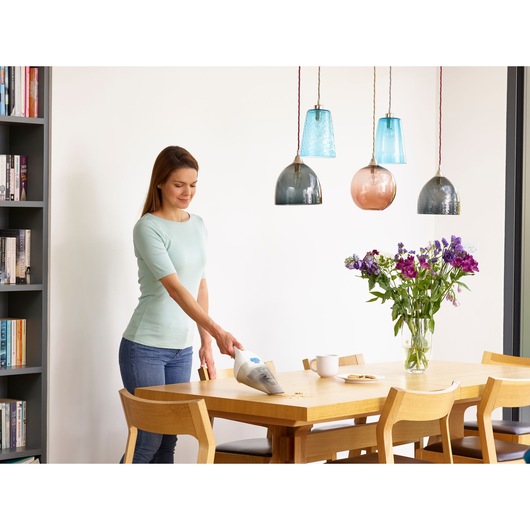 BLACKDECKER NVC115JL handheld vacuum cleaning crumbs from wooden dining table near vase with flowers.