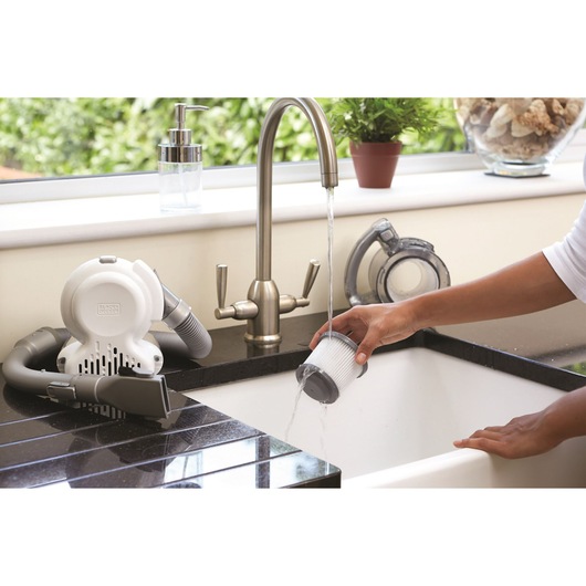 BLACK+DECKER PD1420LP handheld vacuum filter being washed under a kitchen faucet, vacuum on black countertop.