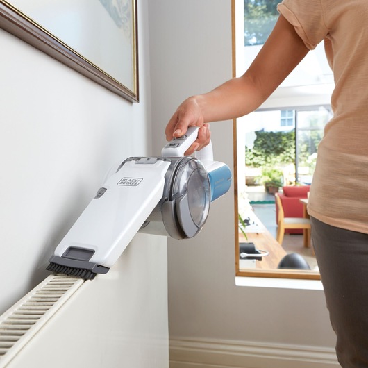 BLACK+DECKER PV1020L handheld vacuum cleaner with brush attachment cleaning a radiator in a bright living room.