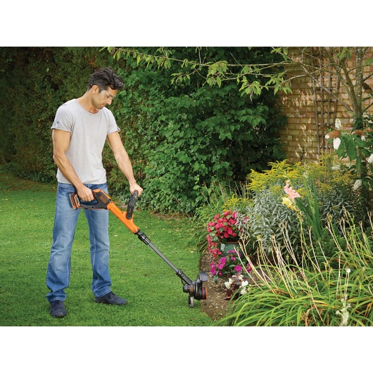 BLACKDECKER STC1820PCB grass trimmer used in garden next to flowerbed, orange and black cordless tool.
