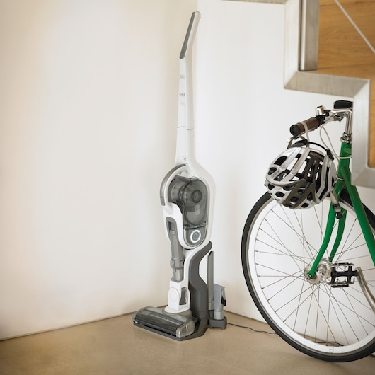 BLACKDECKER SVFV3250L cordless stick vacuum in white and gray on charging base next to green bicycle with helmet.