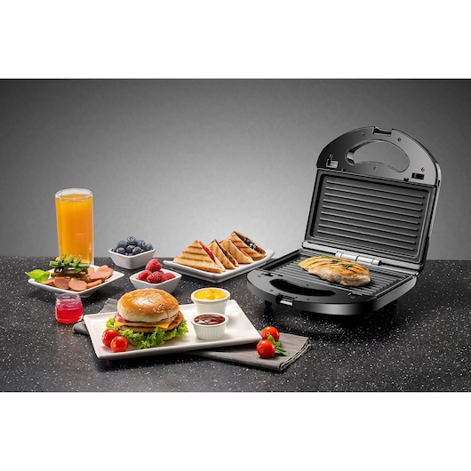 BLACKDECKER TS2130 sandwich maker on countertop with grilled chicken, burger, sandwiches, juice, and assorted fruits.