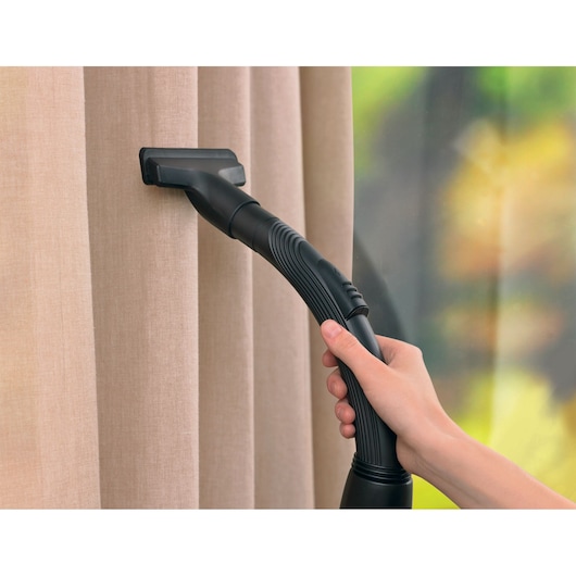 BLACKDECKER vacuum cleaner V01850-B5 cleaning beige curtains with a hand holding the black hose.