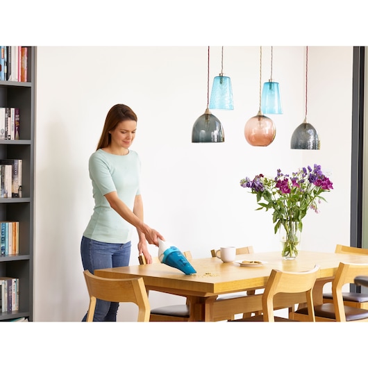 A person using a handheld BLACK+DECKER vacuum cleaner (model WDC115WA) to clean crumbs from a wooden dining table in a bright, modern room. There are flowers in a vase and a cup on the table.