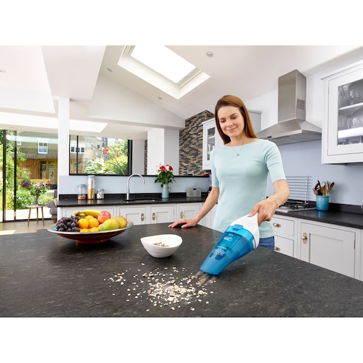 A person using a BLACK+DECKER handheld vacuum cleaner (model WDC115WA) to clean spilled oats on a kitchen countertop. A bowl with oats and a plate of fruit are also on the counter. The kitchen is modern and bright with large windows and a skylight.