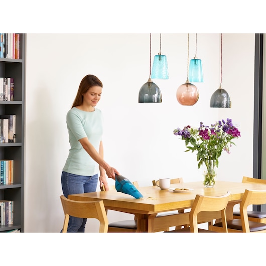 Person using a BLACK+DECKER handheld vacuum cleaner on a wooden dining table with some crumbs, flowers in a vase, cups, and plates, in a modern dining room setting.