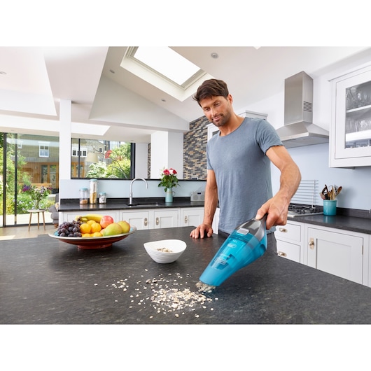 A person using a BLACK+DECKER handheld vacuum cleaner (WDC215WA) to clean spilled oats on a kitchen countertop. There is a bowl of oats and a plate of assorted fruit visible on the counter. The kitchen is modern with large windows and white cabinets.
