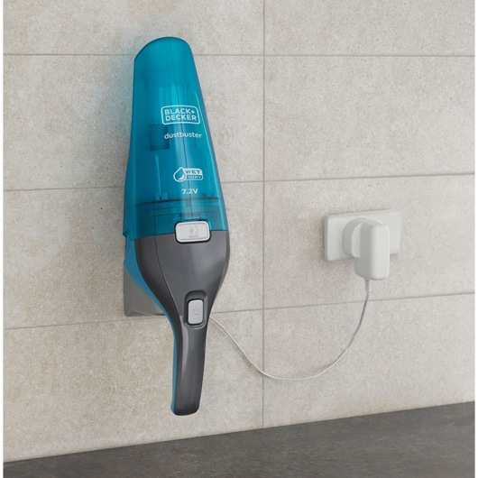 A BLACK+DECKER Dustbuster WDC215WA handheld vacuum cleaner mounted on a wall, charging with a cord plugged into a white socket. The vacuum has a blue and gray design and is suitable for wet and dry cleaning.