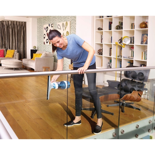 A person is using a BLACK+DECKER WW100 window and glass cleaner to clean a glass railing inside a modern living room with wooden flooring, shelves, and comfortable seating.