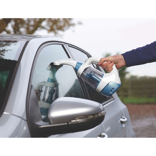 A person using a BLACK+DECKER WW100 handheld cleaning device to remove water from the window of a silver car.