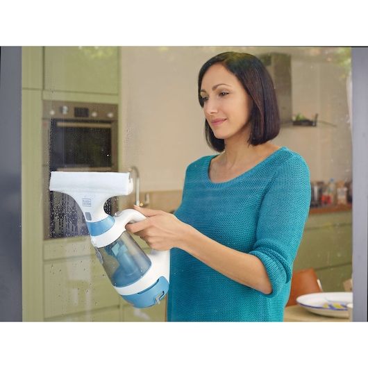A person uses a BLACK+DECKER window vacuum cleaner (model WW100) to clean a glass surface in a kitchen setting. The device is white and blue, and visible water streaks are being removed from the glass.