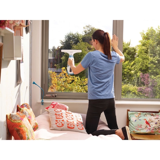 A person kneels on a bed and uses a BLACK+DECKER window vacuum cleaner (model WW100) to clean a large window in a well-lit room with a view of trees outside.