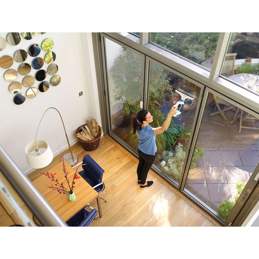 A person is using a BLACK+DECKER WW100 window cleaning device to clean large glass sliding doors in a modern home. The person stands indoors on wooden flooring near a table, chairs, and a floor lamp. The view outside shows a garden patio with plants and outdoor furniture.