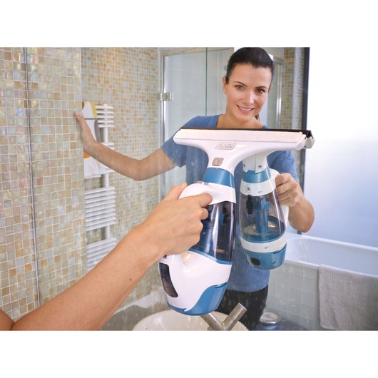 A person using a BLACK+DECKER WW100 window vac to clean a bathroom mirror. The person's face is blurred for privacy. The device is white and blue, and the bathroom has a tiled wall and a sink visible below.