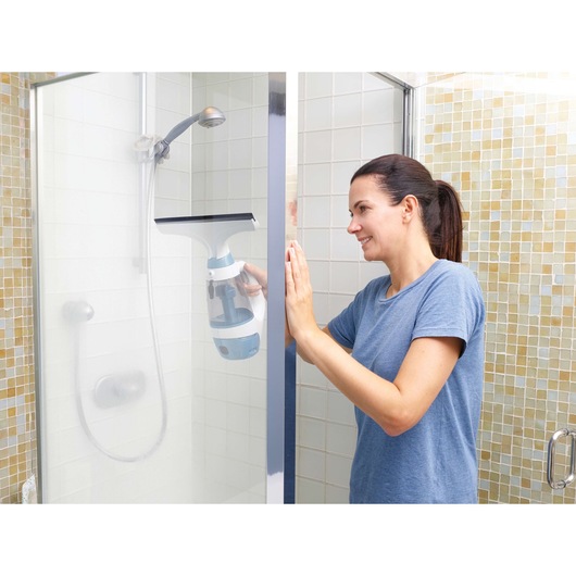A person is cleaning a glass shower door with a BLACK+DECKER window vacuum cleaner inside a bathroom. The person is holding the device against the glass, and the shower area is visible in the background.
