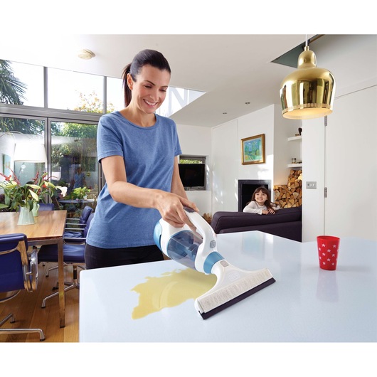 A person uses a BLACK+DECKER WW100 handheld wet vacuum cleaner to clean a liquid spill from a table in a modern, well-lit kitchen and living room.