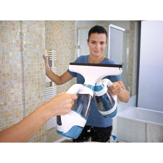 A person cleaning a bathroom mirror with a BLACK+DECKER WW100 handheld window vacuum cleaner. The individual's face is blurred for privacy, and the bathroom has light-colored mosaic tiles.