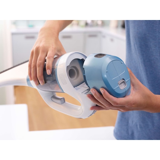 Close-up view of a person removing or attaching the water tank to a BLACK+DECKER handheld cleaning device, in a modern kitchen setting.