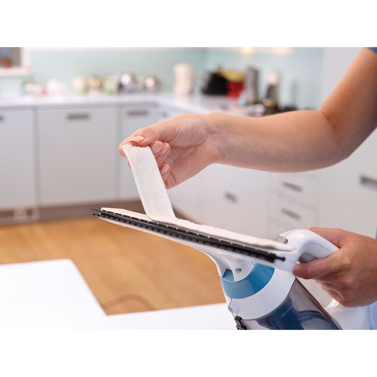 A person is holding a BLACK+DECKER window vacuum cleaner in a kitchen and attaching a cleaning pad to the device.