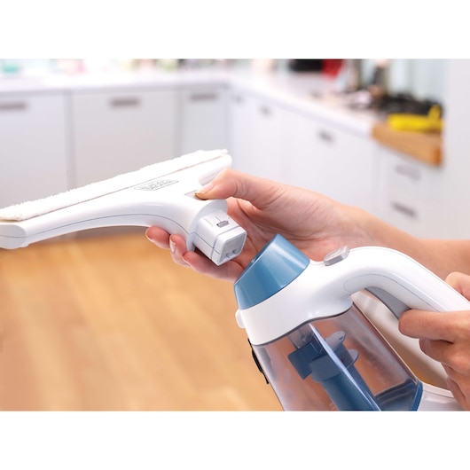 A close-up of hands assembling a Black+Decker window cleaning appliance, with a removable cleaning head and water tank, in a modern kitchen setting.