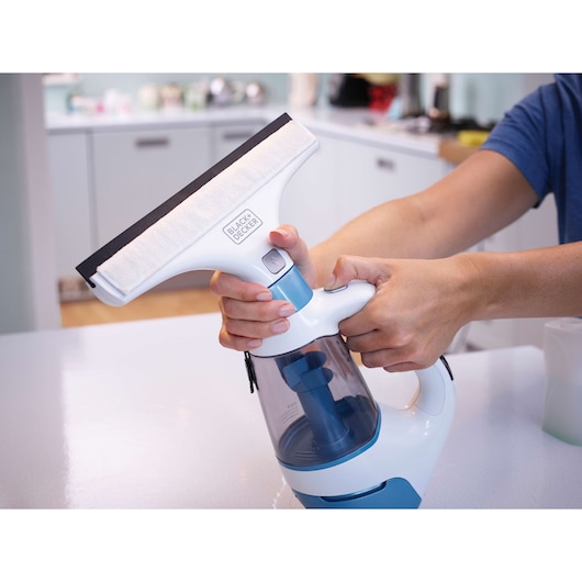 A person is using a BLACK+DECKER window vacuum cleaner in a kitchen setting. The device has a squeegee head and a transparent container.