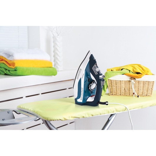 BLACK+DECKER X2150 steam iron on yellow ironing board with folded towels and laundry basket.