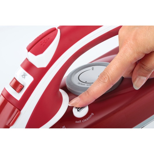 A close-up of a red and white steam iron with a hand pressing the self-cleaning button near the temperature control dial.